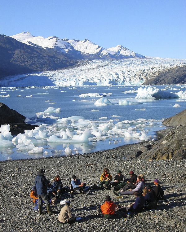 Circle of students gather around an instructor for a lesson in Patagonia with icebergs floating in the water behind. 