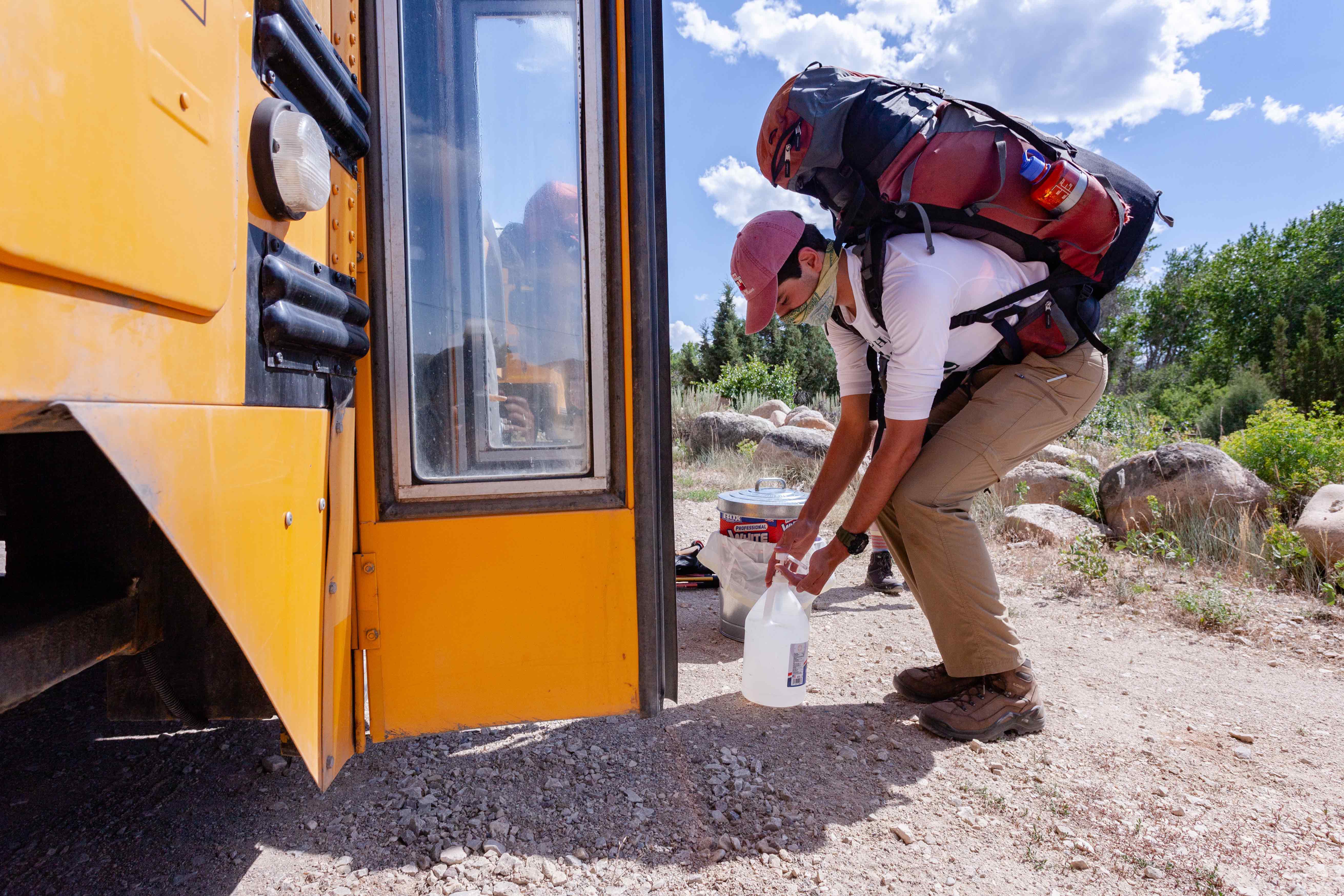 A student wearing a backpacking pack and face mask bends down to dispense some hand sanitizer before getting on a school bus.